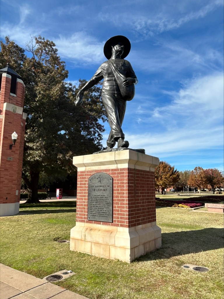 A larger-than-life bronze statue of a man on a red brick plinth in a wide-brimmed hat and pioneer garb spreading seeds.